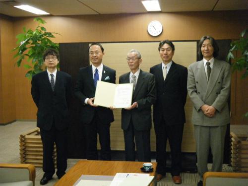 スーツを着た5人の男性が並び、左から2番目の市長と関係者の男性が一緒に協定書を持って記念撮影をしている写真