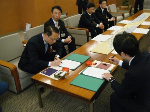 中央に置かれたテーブルの左側に座っている市長と、右側に座っている男性がそれぞれの書類に記入をしている写真
