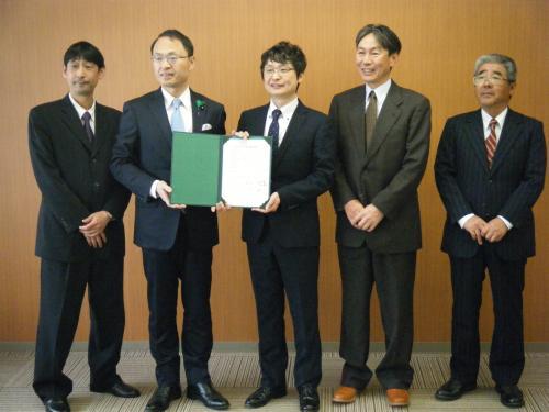 スーツを着た男性5名が横一列に並び、市長と関係者の男性が一緒に協定書を持って記念撮影をしている写真
