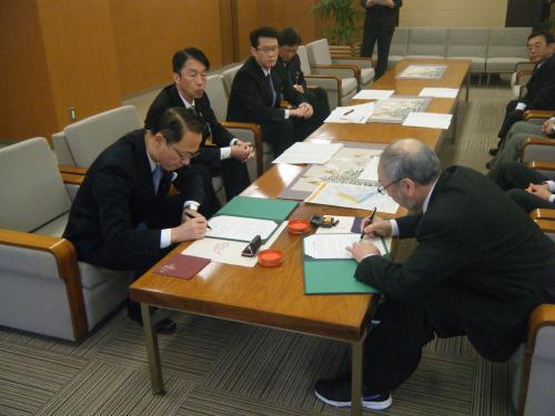 中央に置かれたテーブルの手前側に座っている市長と関係者の男性がそれぞれの書類に記入をしている写真