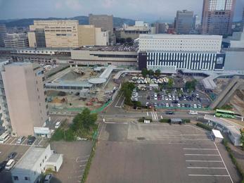 平成24年8月の工事進捗、北西から撮影した写真。左手前に茶色いビルが建っていて、奥に工事中の駐車場が見えている。