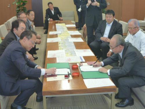 テーブルの左側に座っている市長が書類に判を押し、右側に座っている男性が書類に記入をしている写真