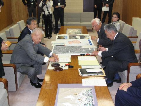 テーブルに資料などが広げられ、その横で市長と関係者の男性がそれぞれの書類に判を押している写真