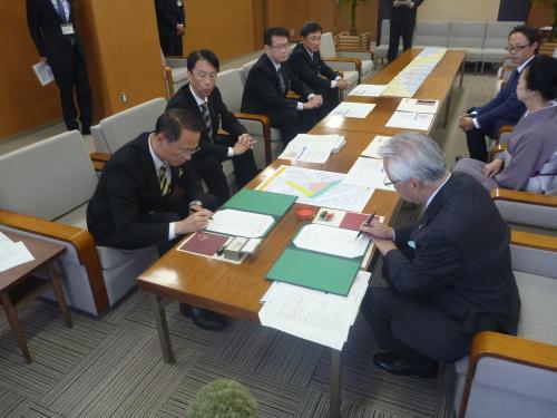 中央のテーブルを挟んで座っている関係者の人達の、手前に座っている男性2名がそれぞれ書面に記入をしている写真