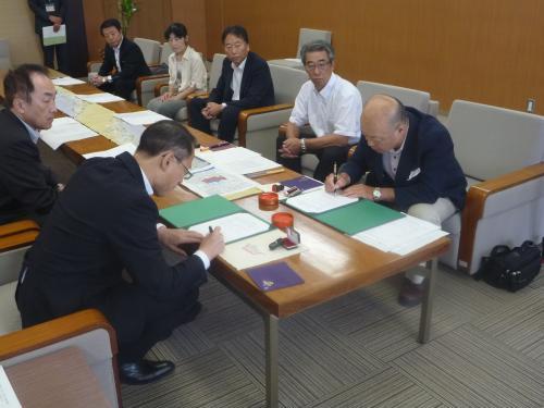 中央に置かれたテーブルを挟んで両側に関係者達が座り、手前側に座っている市長と関係者の男性が書類に記入をしている写真