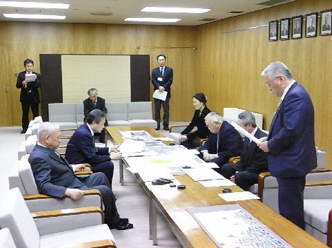 山出金沢市長と男性が中央に置かれたテーブルの左側に座り、右側に3名の関係者が椅子に座り、1人の男性が手に書類を持って立っており、協定に向けて話し合いが行われている写真