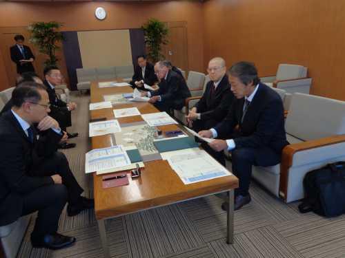中央のテーブルを挟んで左側に3名の男性、右側に5名の男性が座り、話し合いが行われている様子の写真