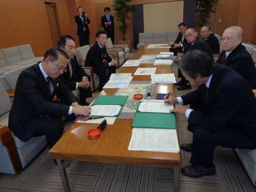 テーブル手前に座っている左側の市長が書面に判を押し、右側の彦三五番丁上組親交会の関係者の男性が書面に記入をしている写真