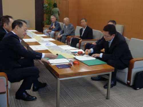 中央に置かれたテーブルの手前画に座っている市長と関係者の男性がそれぞれの書類に判を付こうとしている写真