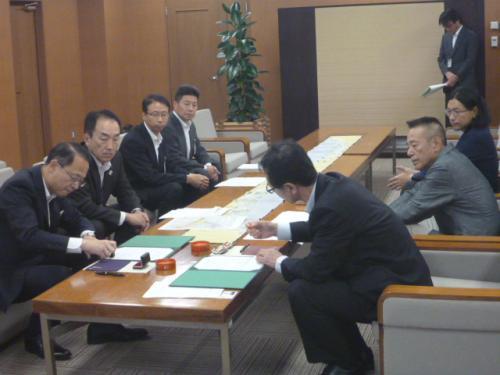 中央に置かれたテーブルの手前側に座っている男性2名が書類に判を押そおとしている写真