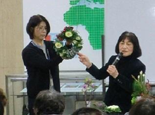 左の女性が持つ花のリースの説明を右の女性がマイクを持ち説明をしている写真