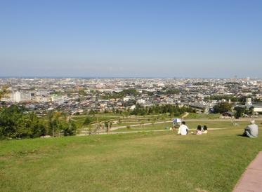 公園の丘の上から斜面になっている芝生と芝生に座っている人々と建物などの風景を撮影した写真