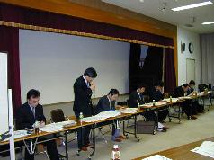 会場の前方に長テーブルを一列にして設置された椅子に2名ずつ男性が座り、左から2番目の男性が立ってマイクを片手に持ち話しをしている写真