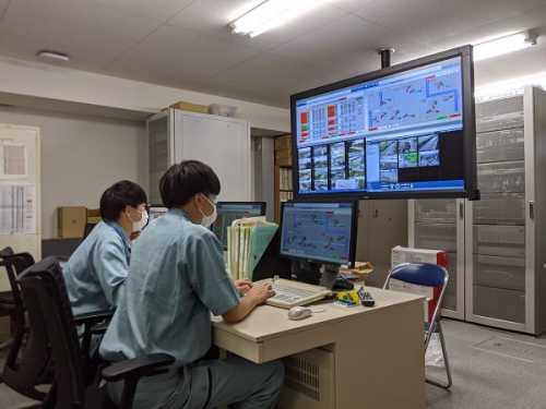 作業着を着た男性職員2名が並んで座り、パソコンや前方の大きな液晶の画面を見ている写真