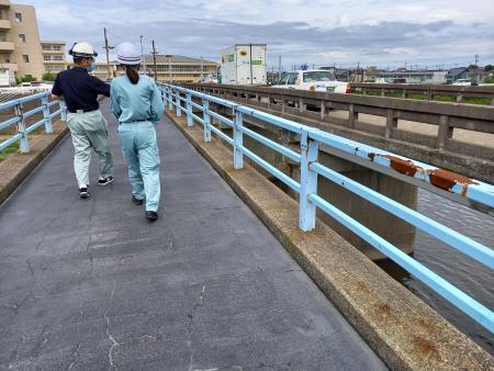 ヘルメットをかぶり、作業着を着た2名の職員が、トラックやタクシーが通っている道路橋の横を歩いている後姿の写真