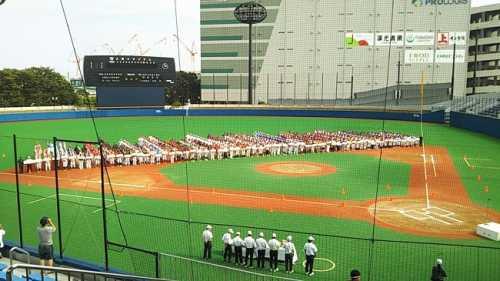 野球場で内野と外野の間あたりからホームベース方向に向かって整列して並んでいる大勢の野球チームと参加選手を観客席から撮影した写真