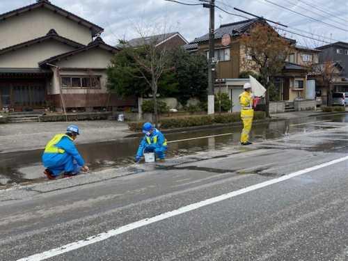 濡れた道路で旗を持って立っている黄色の作業着を着た男性と、青色の作業着を着た男性2人がしゃがんで作業をしている写真