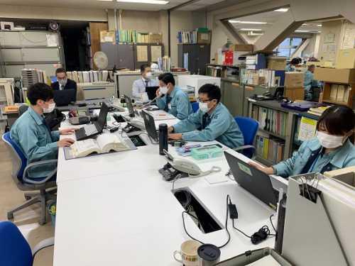 きれいに整理された白い長机に作業服をきた男女4名がパソコンに向かって座り作業している写真