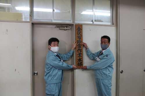 作業服を着ている男性2名が入り口の右側の壁に「金沢市除雪作業本部」と書かれた木製の看板と取り付けている写真