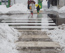 傘をさした通行人が歩く横断歩道の左右端に、雪が山積みになっていて、手前の山積みの雪の間に薄く雪と水が混じったもので覆われている写真