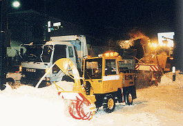 道路には高く雪が積もっており、夜間照明が付けられた中、トラックの横で小型除雪車が除雪作業をしている写真