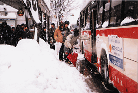 バス停前に大量の雪が積み上げられ、バスが車道側溝部から離れた場所に停車している為、乗り込もむ乗客がバスの側面に手を伸ばし乗車するのに手間取っている様子の写真