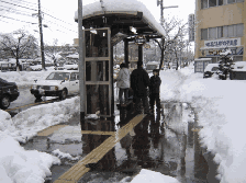 バス停の屋根に雪が積もり、市民が待つバス停の周りが無雪化されている写真