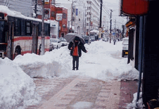 傘をさした通行人が、歩道に積もった雪の上を慎重に歩いている写真