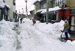 住宅前の道路に沢山の雪が降り積もり、住民の方々がスコップで道路の雪かきをしており、高齢の女性がスコップを持って、道路脇に立っている除雪作業の様子の写真