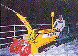 夜間に男性が、黄色い車体の小型除雪機を操作し、歩道に降り積もった雪の除雪作業を行っている写真