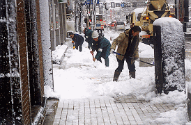 沿道沿いの歩道に積もった雪を、スコップを使って手作業で人々が除雪作業をしている写真