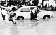 冠水した道路にある車の運転席のドアを開けて、車を押しているような男性と、膝あたりまできている水の中、傘をさして歩いている女性の白黒写真