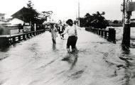 川の水が橋の上や道路に流れ込む中、橋の上を歩く数名の男女の様子が写っている白黒写真