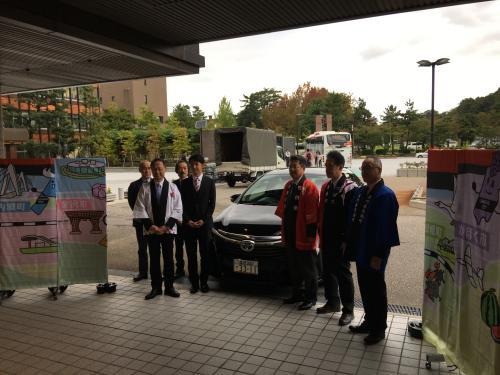 図柄入り金沢ナンバーの車を中心に法被を着た男性4名とスーツ姿の男性3名がお披露目会で記念撮影をしている写真