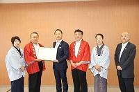 赤の法被の男性の隣に割烹着姿のおかみさんの計4名と関係者2名で締結書を持ち記念撮影をしている写真