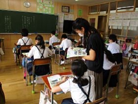 教室で生徒達が席に着き、机上の教科書などを見ており、後方に座っている女の子の右側に立っている黒い服を着た女性が教科書を見ている写真