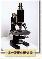 机の上に顕微鏡が置かれていて「博士愛用の顕微鏡」と文字が入っているモノクロ写真