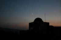 夕方ごろの天文学習棟の隣にうっすら白い三日月が昇る様子の写真