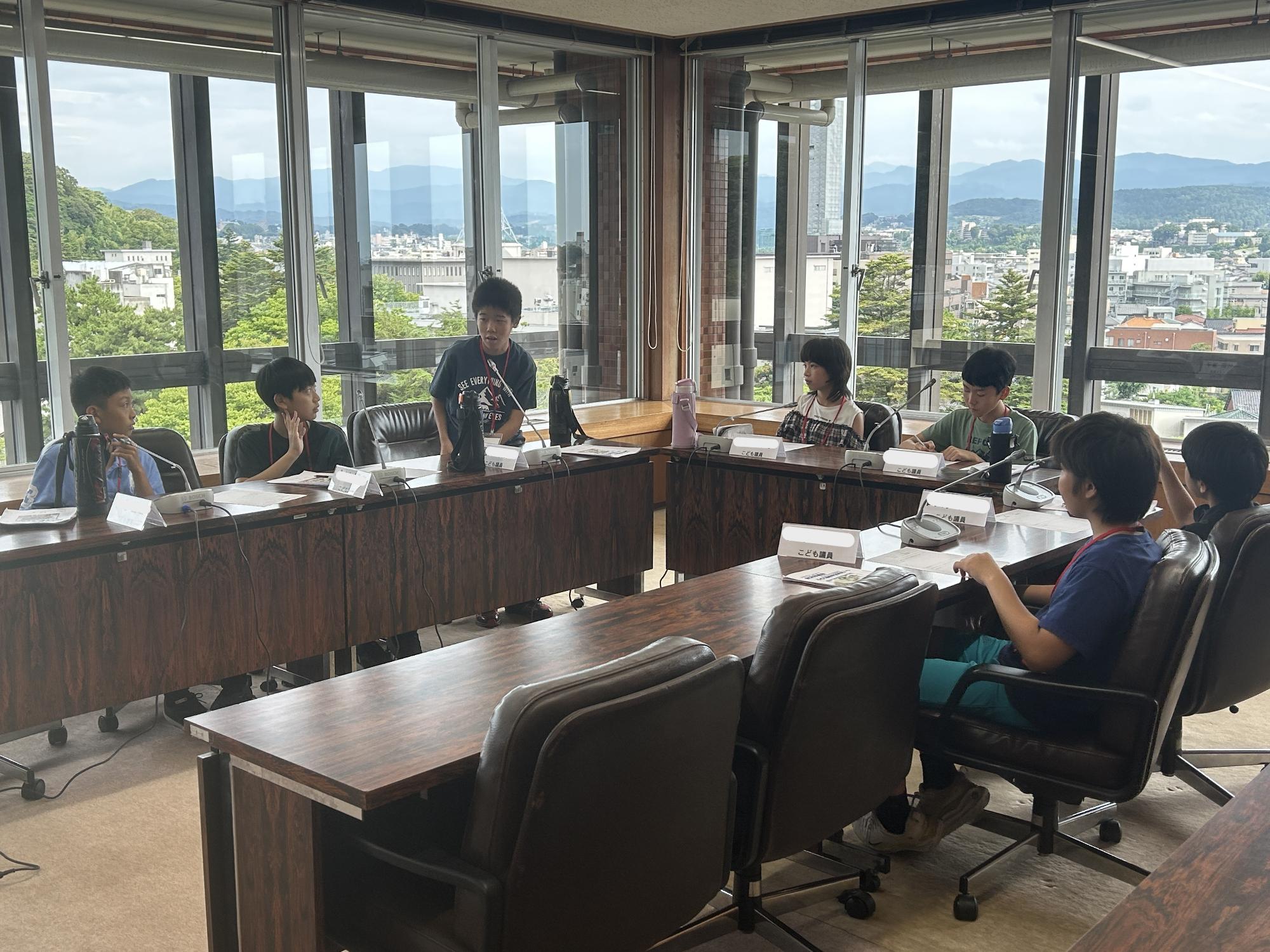 会議室にコの字に設置された長机の椅子に座っている子供たちが議員体験を行っている様子の写真